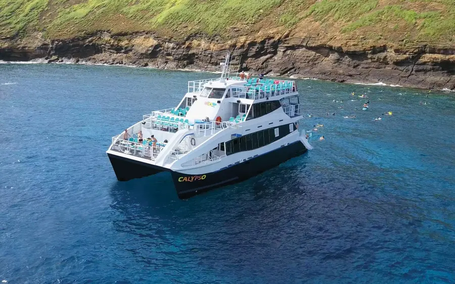 Un gran catamarán blanco de tres cubiertas sobre aguas azules bajo acantilados verdes