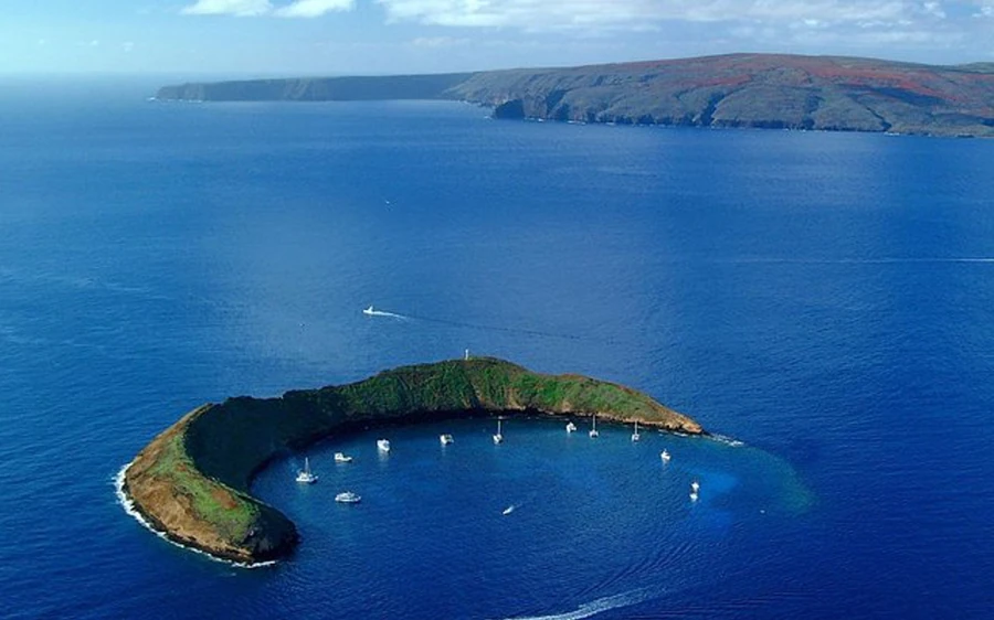 El islote en forma de media luna visto desde el aire con pequeñas embarcaciones blancas fondeadas dentro de la bahía